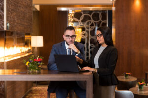 White man, white woman dressed in business clothes looking at computer in fancy lobby.