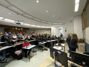 Learners sit in tiered seating in an Algonquin College Ottawa campus classroom. Students jot down notes in notebooks and on laptops while the President delivers his answers to questions posed during the press conference.