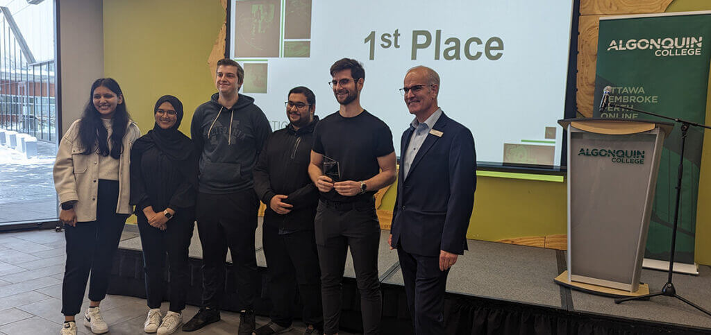Six people stand smiling in front of a projector screen with the words "1st Place" written on it.