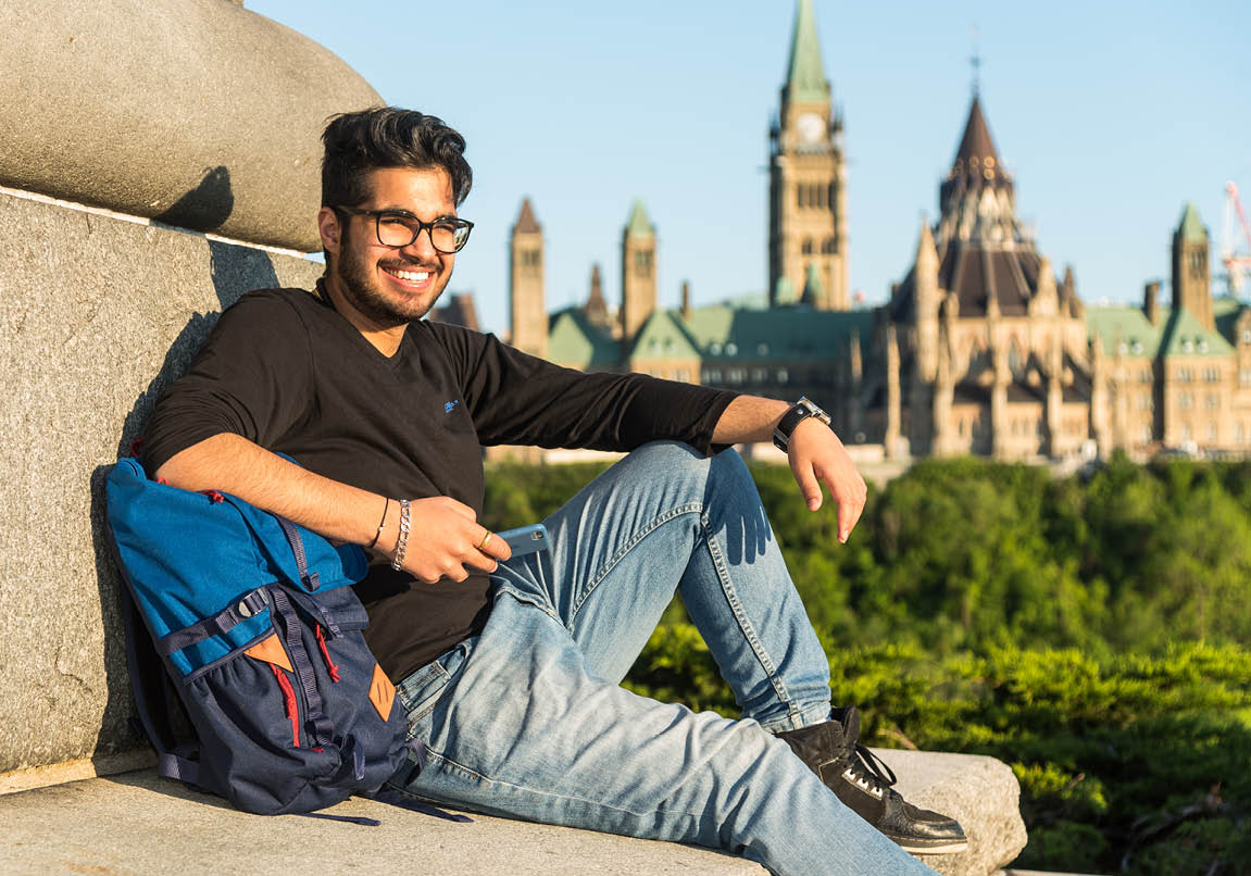 Photo of an international student smiling and sitting with their backpack outside with the parliment buildings in the background. 