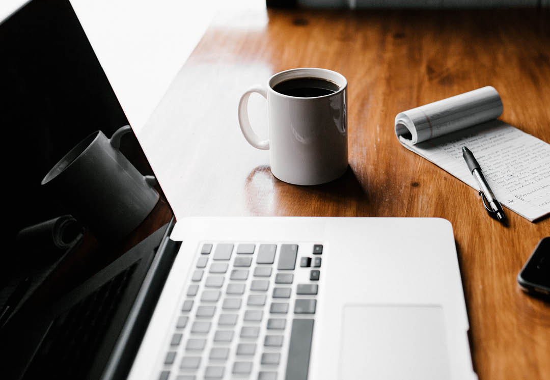 Photo of a desk setup with a laptop, coffee, and notepad.