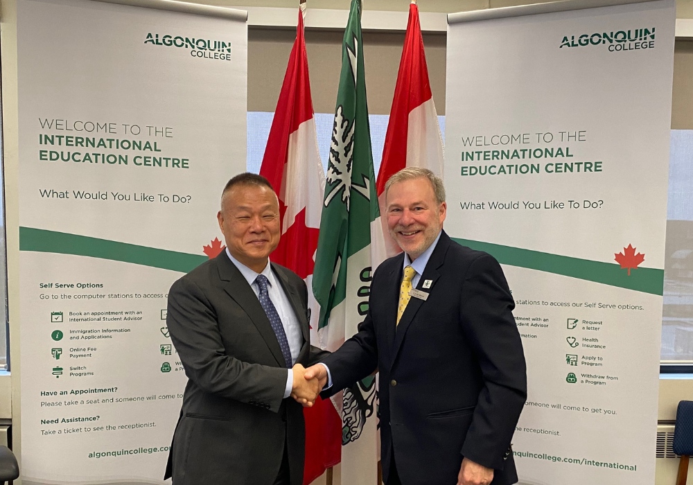 Algonquin College President &amp; CEO, Claude Brulé, shakes hands with CDI College President & CEO, Dr. Peter Chung, in celebration of the partnership agreement's official signing. The pair stand in front the Algonquin College and Canadian flags alongside Algonquin College banners that read "Welcome to the International Education Centre".
