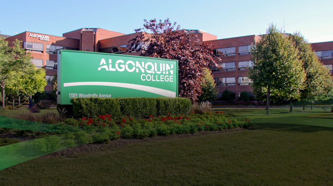 Photo of Algonquin College's B Building from the intersection of Woodroffe Avenue and College Avenue. A green sign with the Algonquin College logo and address stands in front of the building in summer time with red flowers planted around it. Several trees are in the foreground between the sign and the building.