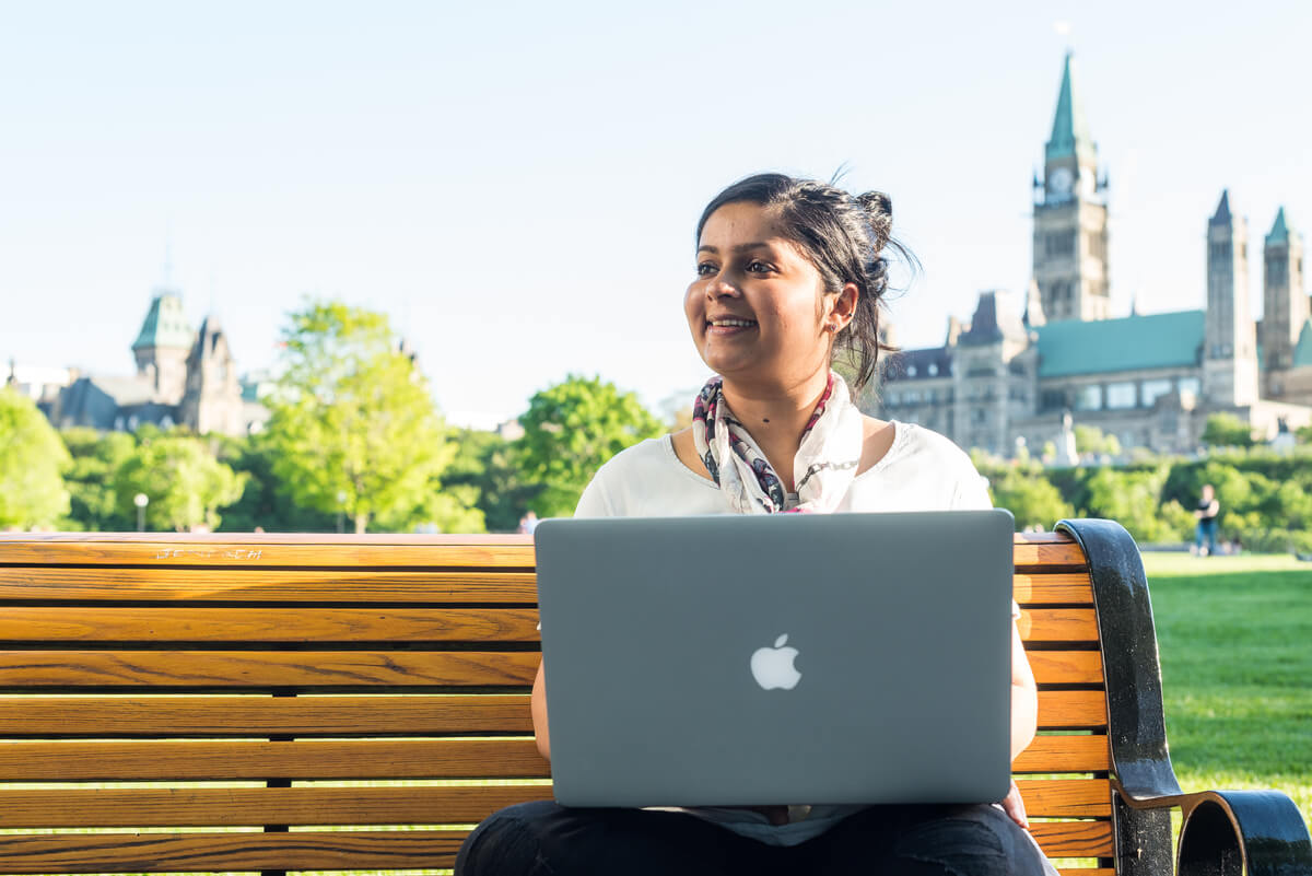 A woman sitting on a bench outside using a laptop. The Canadian parliament building is behind her.