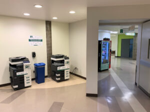 Image of one Konica Minolta colour printer and one Konica Minolta black and white printer on the second floor of B building in the hallway near room B261 and around the corner from the link to ACCE building.
