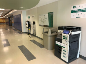 Image of a Konica Minolta black and white printer on the second floor of T building in the hallway outside of the Bits N' Bytes cafe.