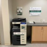 Image of a Konica Minolta black and white printer on the third floor of the Pembroke campus located beside the elevators and across the hallway from room 338.